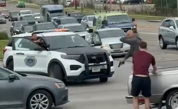 Agente de policía con arma en la ventanilla de una patrulla blanca. Hombre con pistola en la mano. Otro hombre de espaldas. Coches en una calle con tráfico