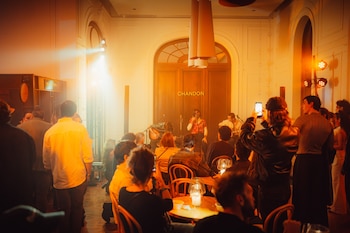 Un grupo musical actúa en un escenario iluminado por luces cálidas, rodeado de asistentes. Se aprecian luminarias colgantes y una puerta con 'CHANDON' al fondo