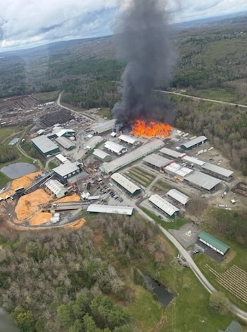 Vista aérea de un complejo industrial con un gran incendio y denso humo negro elevándose. Hay árboles y paisaje rural alrededor de la instalación