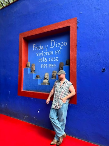 Hombre con gorra y camisa a cuadros sin mangas, jeans y botas, se apoya en pared azul con marco rojo, que muestra bustos de piedra y texto 'Frida y Diego vivieron en esta casa 1929-1954'