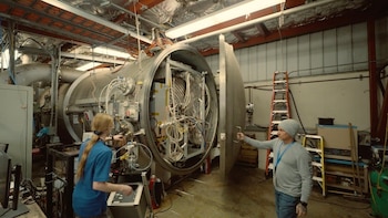 Dos personas en un laboratorio interactúan con un gran propulsor iónico dentro de una cámara cilíndrica de metal, llena de cables y componentes