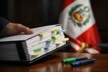 Una mano sujeta un documento encuadernado con pestañas de colores sobre una mesa de madera, junto a dos rotuladores resaltadores y la bandera de Perú al fondo.