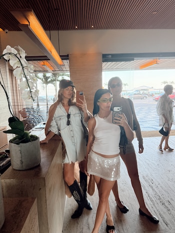 Tres mujeres se toman una foto frente a un espejo en el vestíbulo de un hotel, junto a una maceta con orquídeas blancas