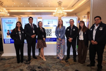 Ocho personas de pie frente a tres pantallas digitales con texto y códigos QR, en un salón con candelabros y alfombra