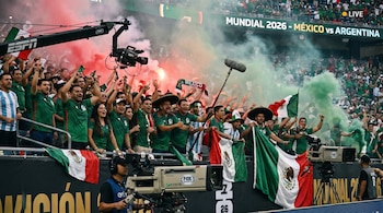 Aficionados mexicanos celebran con banderas y humo de bengalas en un estadio del Mundial 2026. Se observan cámaras de transmisión y una pantalla con el marcador.