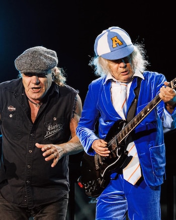 Brian Johnson y Angus Young de AC/DC en el escenario. Johnson usa gorra y camisa oscura; Young viste un traje azul de terciopelo y gorra con la letra 'A', tocando la guitarra