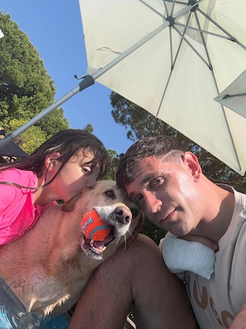 Dibu Martínez, una niña y un perro mojados posan para una selfie en una piscina. La niña besa al perro, que tiene una pelota naranja en la boca, bajo un paraguas blanco