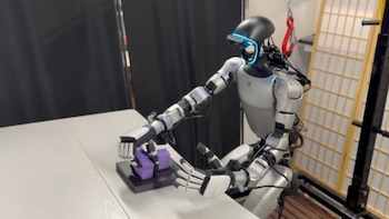 Robot humanoide bípedo de color plata y negro con cabeza negra y visor azul brillante manipulando bloques púrpuras sobre una mesa blanca en un laboratorio