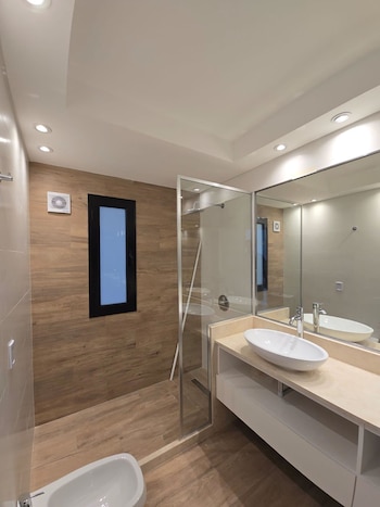 Baño moderno con paredes de azulejos marrones tipo madera y azulejos beige. Cuenta con un lavabo blanco ovalado, espejo grande, ducha de cristal y bidé