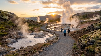 Personas caminan por un camino de grava junto a columnas de vapor y un géiser en erupción en un valle montañoso al atardecer.