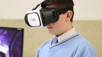 Un niño con camisa, corbata y suéter lleva un visor de realidad virtual sobre los ojos frente a una pantalla