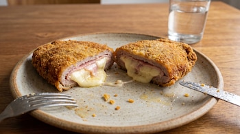 Primer plano de una milanesa rellena de jamón y queso, cortada por la mitad en un plato. El queso fundido se desborda, con cubiertos y un vaso de agua al fondo.