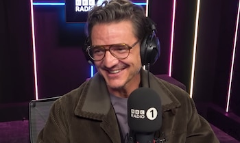 Pedro Pascal sonriendo y riendo, con gafas y auriculares negros, en un estudio de radio. Un micrófono de BBC Radio 1 está en primer plano. Luces de neón en la pared