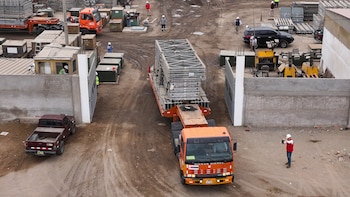 Un camión anaranjado traslada estructuras metálicas de un puente modular al salir de un almacén en un terreno de tierra