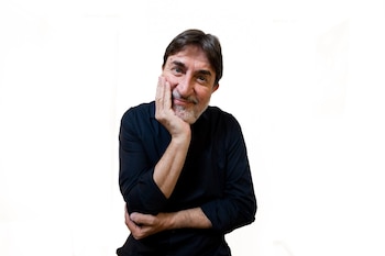 Hombre de cabello oscuro y barba canosa, con camisa negra, apoya la mano en la mejilla sobre un fondo blanco