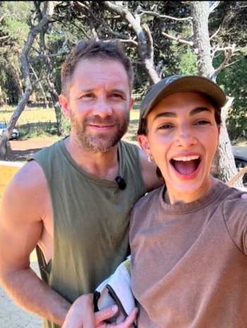Jeremy Camp, un hombre barbudo con camiseta sin mangas verde, sonríe junto a una mujer con gorra verde y camiseta marrón que ríe con la boca abierta. Árboles de fondo