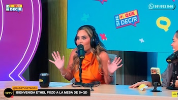 Una mujer con top naranja y auriculares habla por micrófono en una mesa, con un logo de "Sin + Que Decir" al fondo y una taza negra