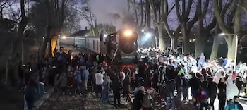 Personas reunidas a los lados de unas vías junto a grandes árboles mientras un tren a vapor con luz encendida avanza entre la multitud