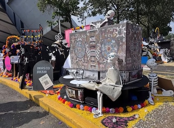 Figuras de esqueletos de papel, una estructura con cintas de película, una lápida decorativa y flores amarillas en el suelo