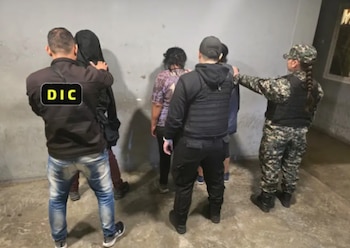 Agentes de policía detienen a varias personas. Uno de los agentes lleva una chaqueta con las letras DIC. Hay tres personas detenidas contra una pared gris