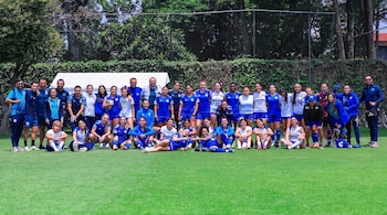 Un grupo grande de mujeres y hombres con uniformes deportivos azules y blancos posa en un campo de césped verde. Hay una valla con vegetación al fondo.