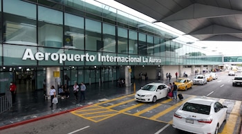 La entrada del Aeropuerto Internacional La Aurora: un edificio moderno con fachada de vidrio, taxis, pasajeros y equipaje bajo una cubierta gris.