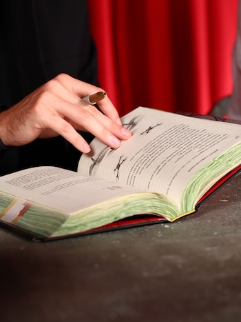 Una mano sostiene un marcador dorado sobre un libro abierto con bordes de páginas verdes, frente a una cortina roja