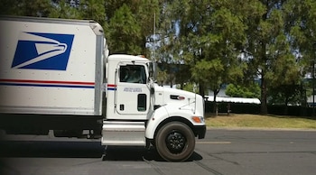 Camión de reparto blanco con el logotipo azul y rojo del Servicio Postal de Estados Unidos, texto "United States Postal Service" en la cabina, camino asfaltado, árboles verdes