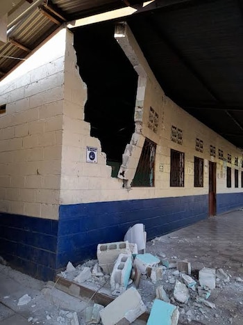 Pared de bloques de hormigón parcialmente destruida, con un gran boquete y escombros en el suelo. El edificio tiene techo de chapa y ventanas con rejas