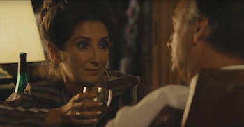 Primer plano de una mujer con cabello oscuro y blusa a rayas, sosteniendo una copa, mirando a un hombre cuyo hombro y parte trasera de la cabeza son visibles. Una lámpara y botella verde al fondo