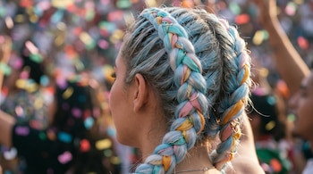 Vista trasera de una persona con trenzas boxer dobles hechas de kanekalon azul, blanco y cintas multicolor. Se observan anillos metálicos y un fondo festivo difuminado.