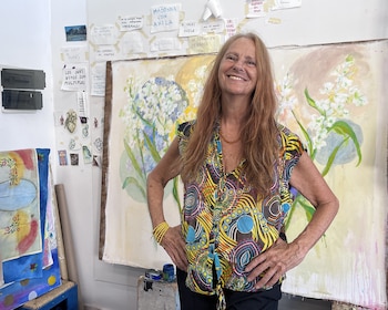 Mujer de cabello castaño rojizo, sonriendo, de pie en un estudio de arte. Detrás, un cuadro de flores blancas y notas manuscritas en la pared. Viste blusa estampada