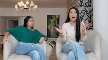 Una mujer de cabello oscuro y otra con rostro difuminado sentadas en sillones. Un perro pequeño está entre ellas. Hay un candelabro y un cuadro.