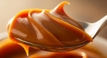 Primer plano de una cuchara metálica con dulce de leche cremoso y espeso, que rebalsa ligeramente el borde, con luz cálida y fondo desenfocado.