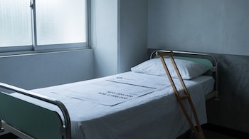 Una cama hospitalaria vacía en una habitación sobria con una factura sobre ella que muestra cifras entre RD$ 200.000 y RD$ 2.000.000, y una muleta.