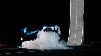 Un automóvil blanco se eleva sobre una densa nube de humo frente a una pista curvada en un entorno nocturno