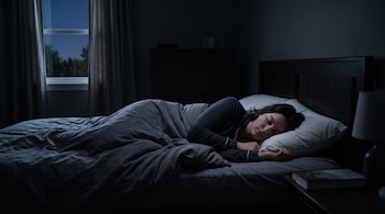 Una mujer duerme en una cama con mantas grises dentro de un dormitorio oscuro con una ventana al fondo.