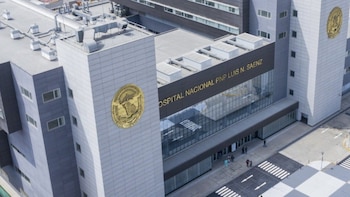Edificio moderno de hospital con fachadas grises y ventanales. Se lee "HOSPITAL NACIONAL PNP LUIS N. SAENZ" en la entrada. Hay emblemas dorados y personas en la acera