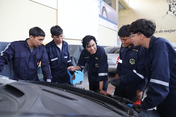 Cinco jóvenes vestidos con overoles azules miran el motor abierto de un coche en un taller, mientras uno de ellos señala un componente del motor
