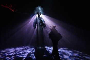 Silueta de una persona tomando una foto con un teléfono a una estatua oscura con tocado de abanico, iluminada por luz púrpura en un espacio oscuro
