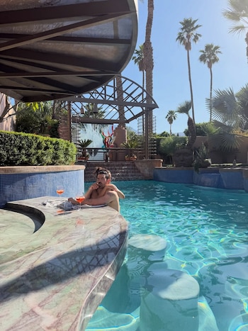 Joe Jonas, sin camiseta y con el pelo recogido, sentado en el bar de una piscina. Dos cócteles naranjas en la barra. Palmeras y cascadas en un día soleado