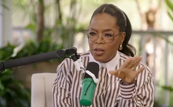 La presentadora Oprah Winfrey con gafas y camisa de rayas marrones y blancas, habla frente a un micrófono verde, gesticulando con la mano derecha