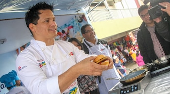 Un cocinero con uniforme blanco sostiene una hamburguesa junto a una sartén, mientras un camarógrafo lo filma y varias personas observan.