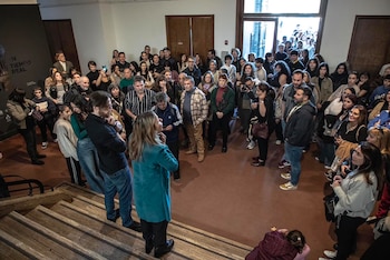 Multitud de personas de pie en una sala interior y escaleras. Una mujer con abrigo turquesa habla a un público diverso. Se ve un banner oscuro con texto