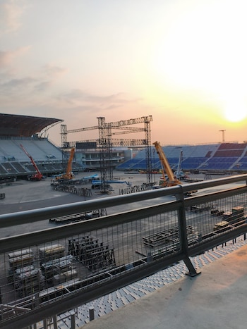 Vista elevada de un estadio donde se monta un gran escenario metálico con grúas y andamios, bajo un cielo anaranjado al atardecer