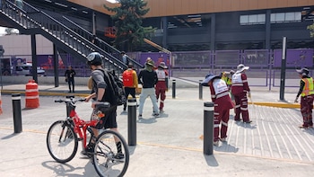 Vista exterior de la estación Metro Universidad con un ciclista y trabajadores de limpieza en uniformes de color guinda, bajo un puente peatonal gris y un edificio púrpura