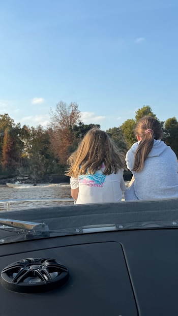 Vista trasera de dos niñas sentadas en un barco, mirando hacia un río con árboles en las orillas bajo un cielo azul. Una usa camisa blanca y la otra una sudadera gris