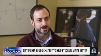 Un hombre blanco con barba y chaqueta burdeos habla en un aula, con una pizarra con dibujos al fondo y un cuadro enmarcado borroso