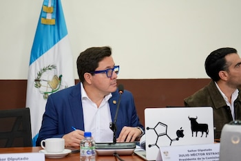 Un hombre con gafas de marco azul habla frente a un micrófono en una mesa donde hay una taza de café y una computadora portátil