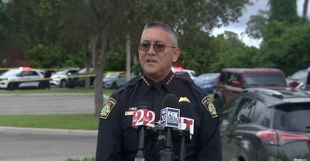 Un oficial de policía con anteojos habla frente a varios micrófonos con vehículos policiales y cinta de seguridad al fondo